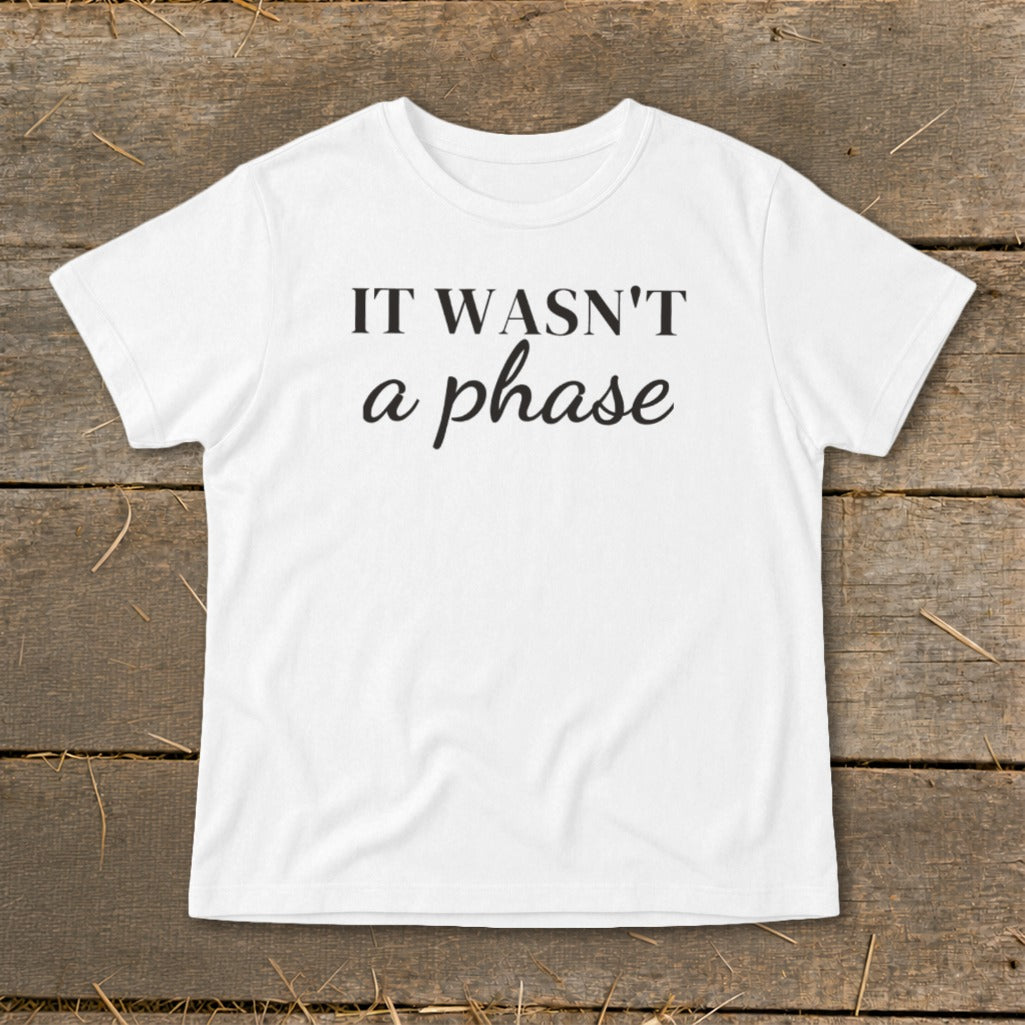 White t-shirt with black text "IT WASN'T a phase" on a wooden background.