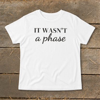 White t-shirt with black text "IT WASN'T a phase" on a wooden background.