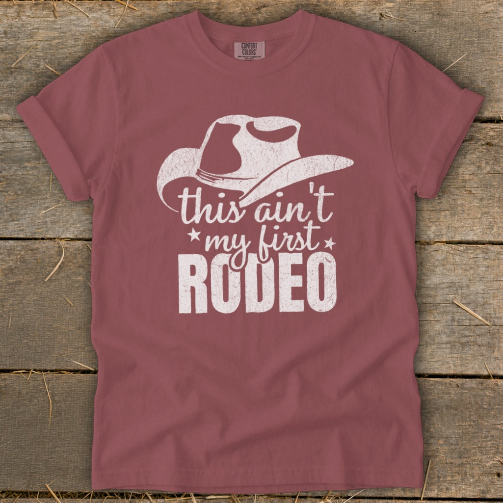 Dusty rose t-shirt with white cowboy hat graphic and "this ain't my first rodeo" text.