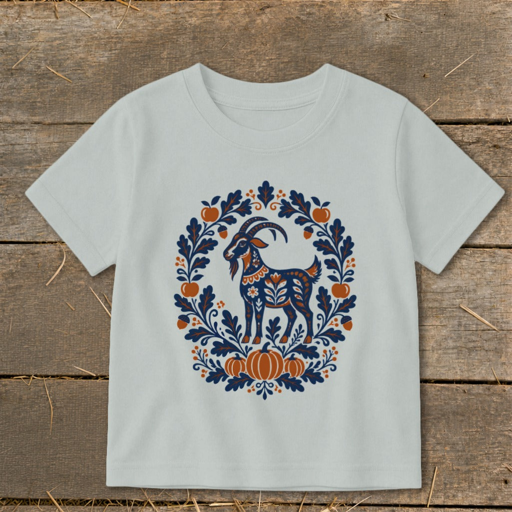 A light blue short-sleeved children's t-shirt features a folk art illustration of a goat framed by autumn leaves, pumpkins, and apples.