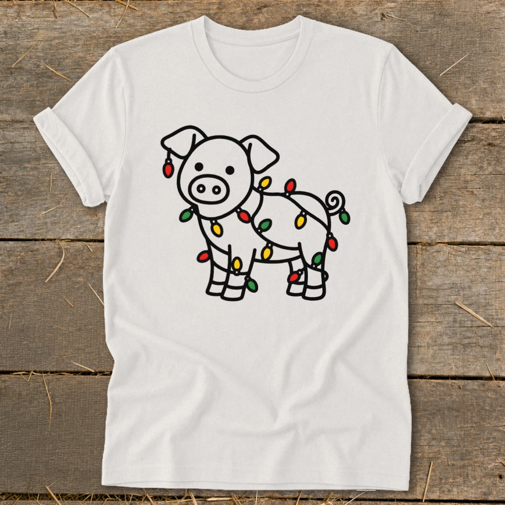 A white t-shirt featuring a cute cartoon pig wrapped in colorful Christmas lights, displayed on a wooden surface.