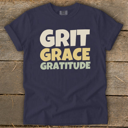Dark blue t-shirt featuring the stacked white and yellow text GRIT GRACE GRATITUDE laid flat on weathered wood.