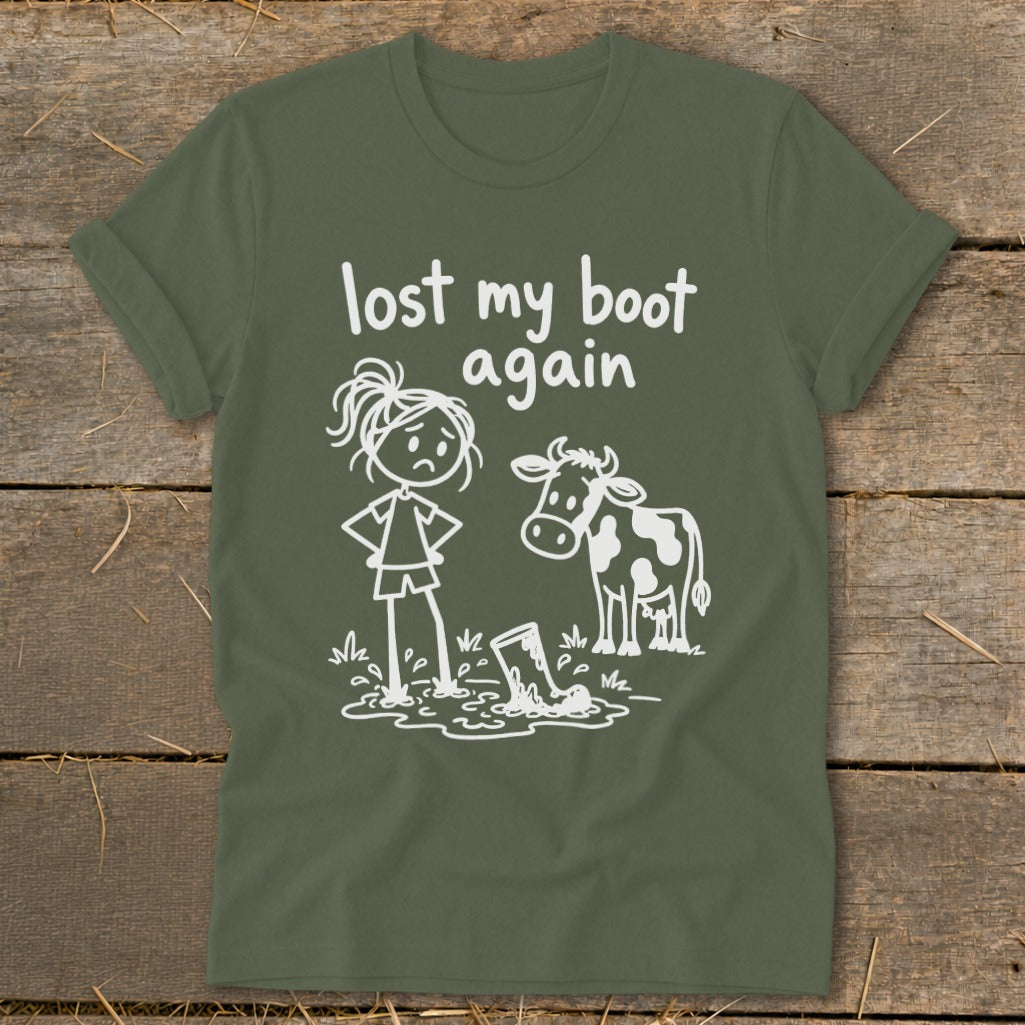 An olive green t-shirt with a white graphic of a sad girl and a cow looking at a boot in a mud puddle, with the text "lost my boot again".