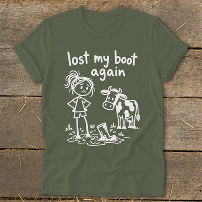 An olive green t-shirt with a white graphic of a sad girl and a cow looking at a boot in a mud puddle, with the text "lost my boot again".