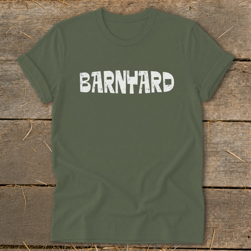 Olive green t-shirt laid flat on a rustic wooden surface with the distressed white word BARNYARD printed across the chest.