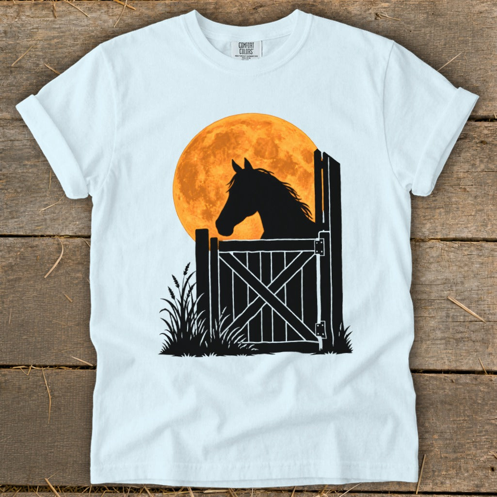 A light blue t-shirt with a graphic of a horse silhouette looking over a stable door against a large orange moon.
