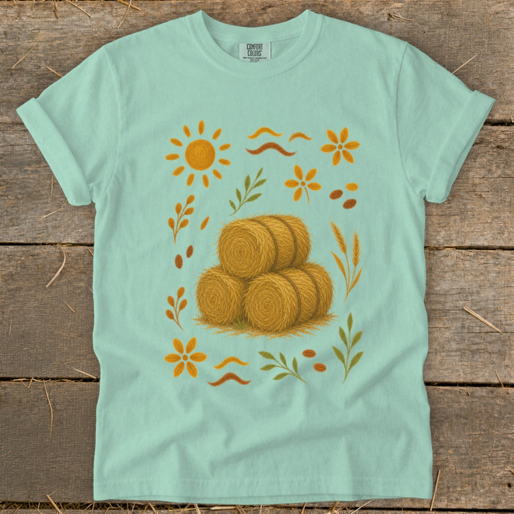 Mint green t-shirt with a stylized autumn harvest graphic featuring hay bales and sun.