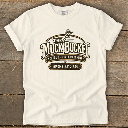 Cream t-shirt with "The Muck Bucket School of Stall Cleaning Opens at 5 AM" graphic.