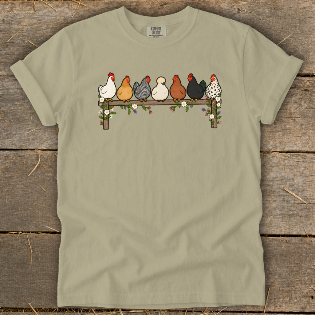 Sage green t-shirt featuring a graphic of seven colorful chickens perched on a flower-covered wooden fence.