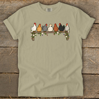 Sage green t-shirt featuring a graphic of seven colorful chickens perched on a flower-covered wooden fence.