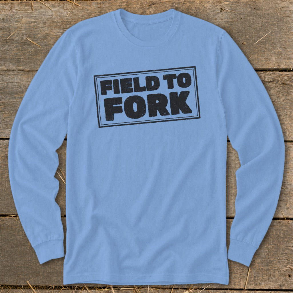 Light blue long-sleeve t-shirt with a distressed black framed graphic reading FIELD TO FORK on a wooden background.