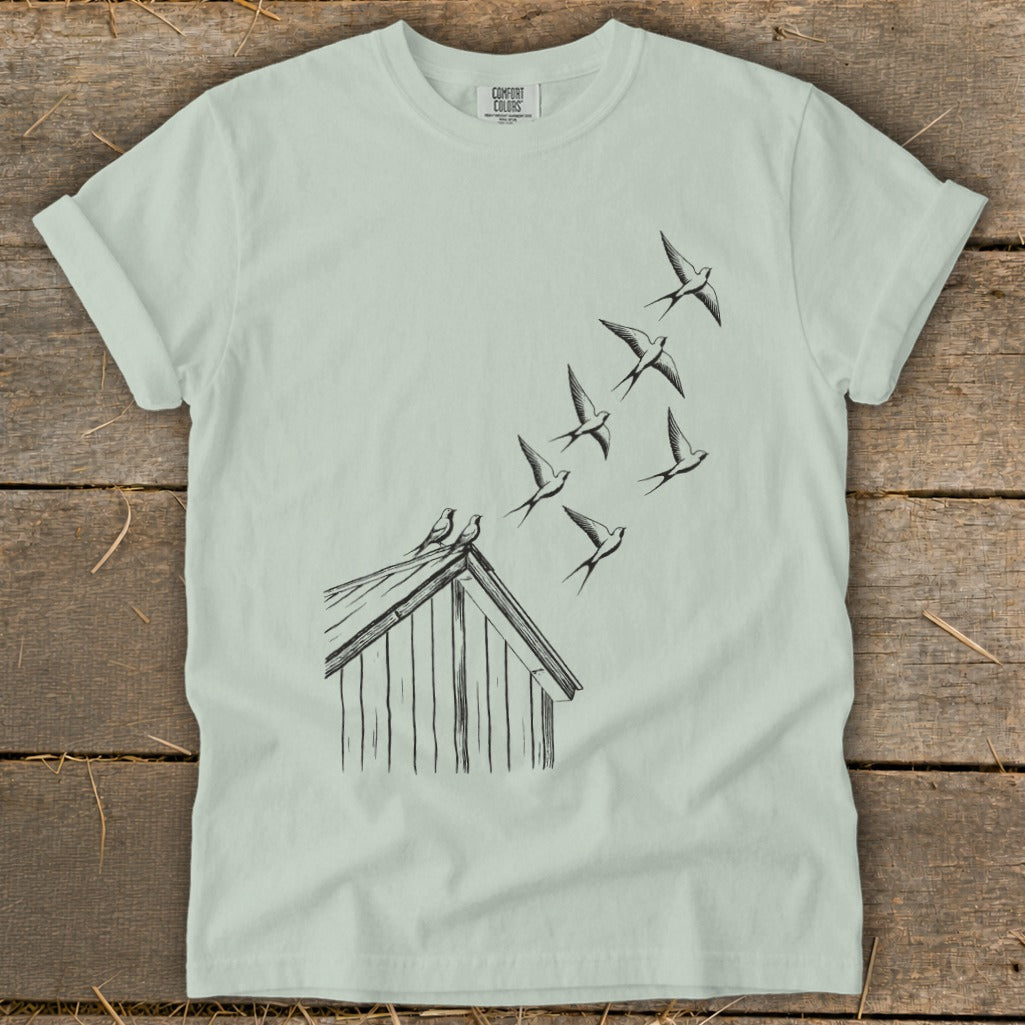A light green t-shirt featuring a black line art graphic of birds flying up from a barn roof.
