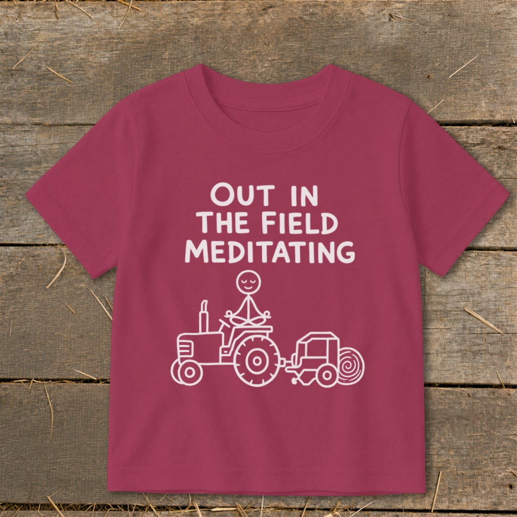 Maroon t-shirt with white text "OUT IN THE FIELD MEDITATING" and stick figure on tractor.