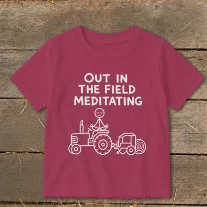 Maroon t-shirt with white text "OUT IN THE FIELD MEDITATING" and stick figure on tractor.