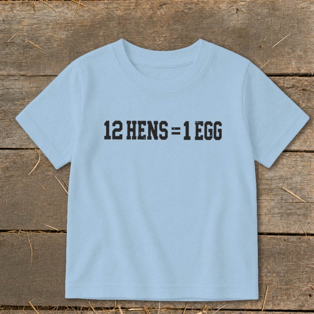A light blue children's t-shirt printed with the black text 12 HENS = 1 EGG, laid flat on a rustic wooden background.