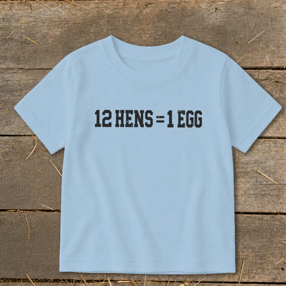 A light blue children's t-shirt printed with the black text 12 HENS = 1 EGG, laid flat on a rustic wooden background.