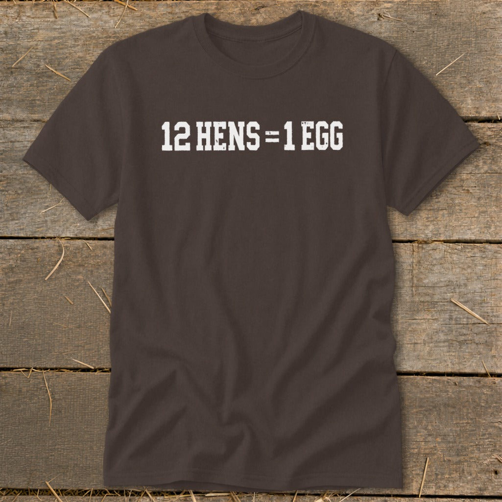 Dark brown short-sleeve t-shirt with white distressed text reading 12 HENS = 1 EGG, laid flat on a rustic wooden surface.