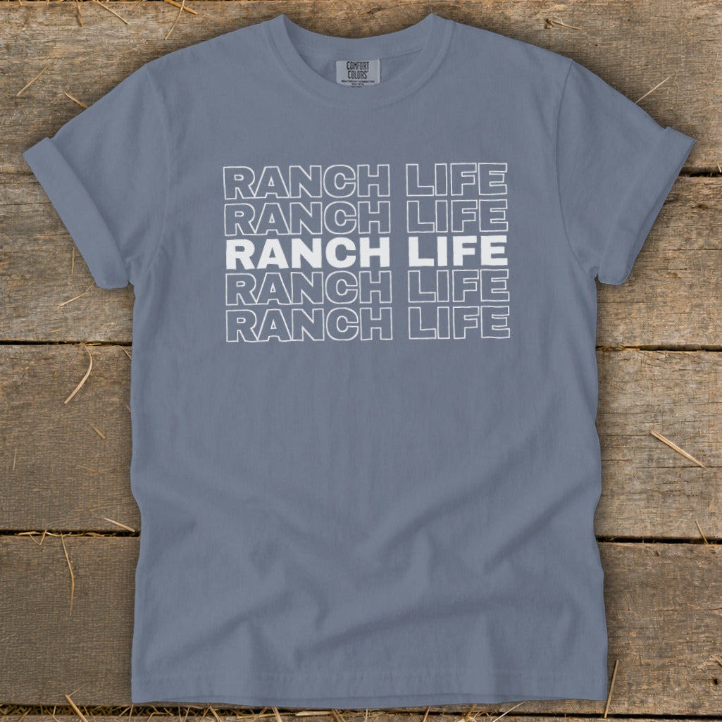 Blue crewneck t-shirt with rolled sleeves featuring repeated white outlined Ranch Life text on a wooden surface.