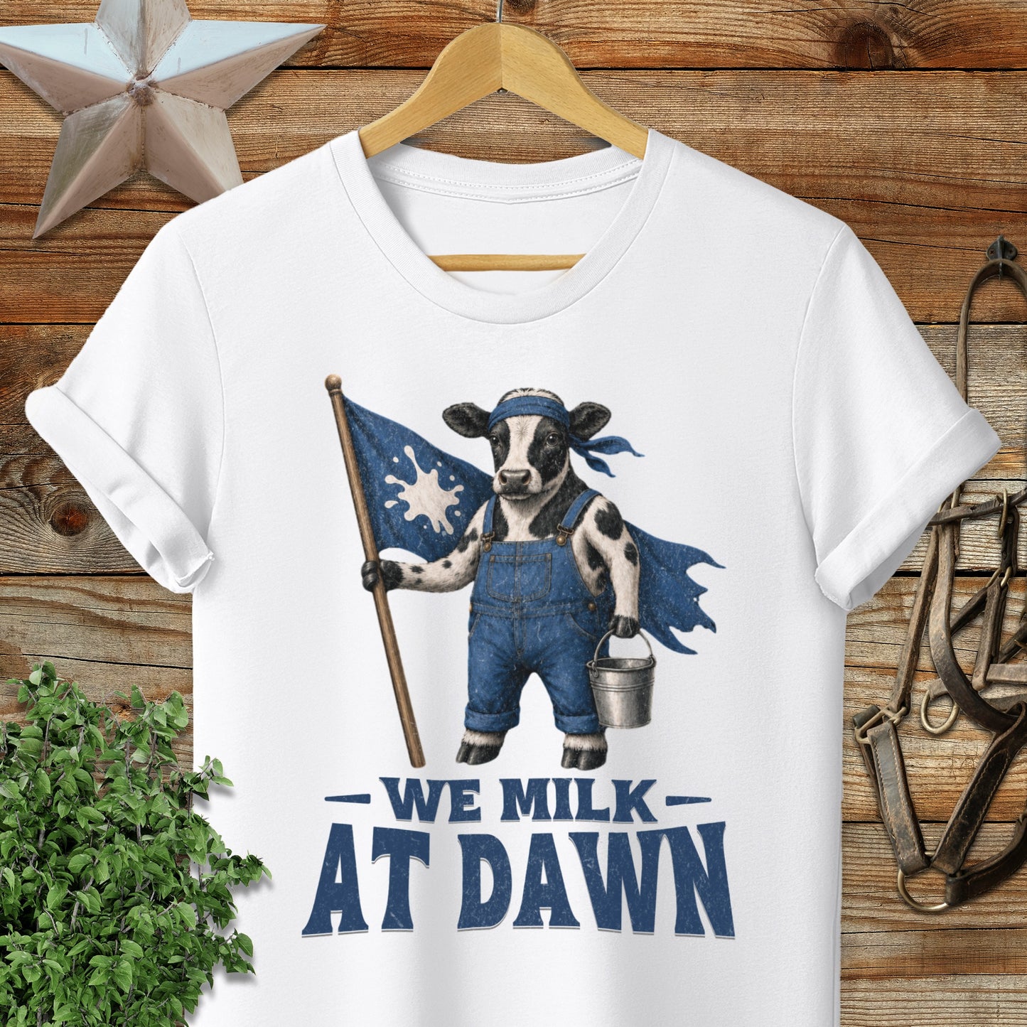 White t-shirt with a cartoon cow holding a flag and bucket, text "WE MILK AT DAWN".