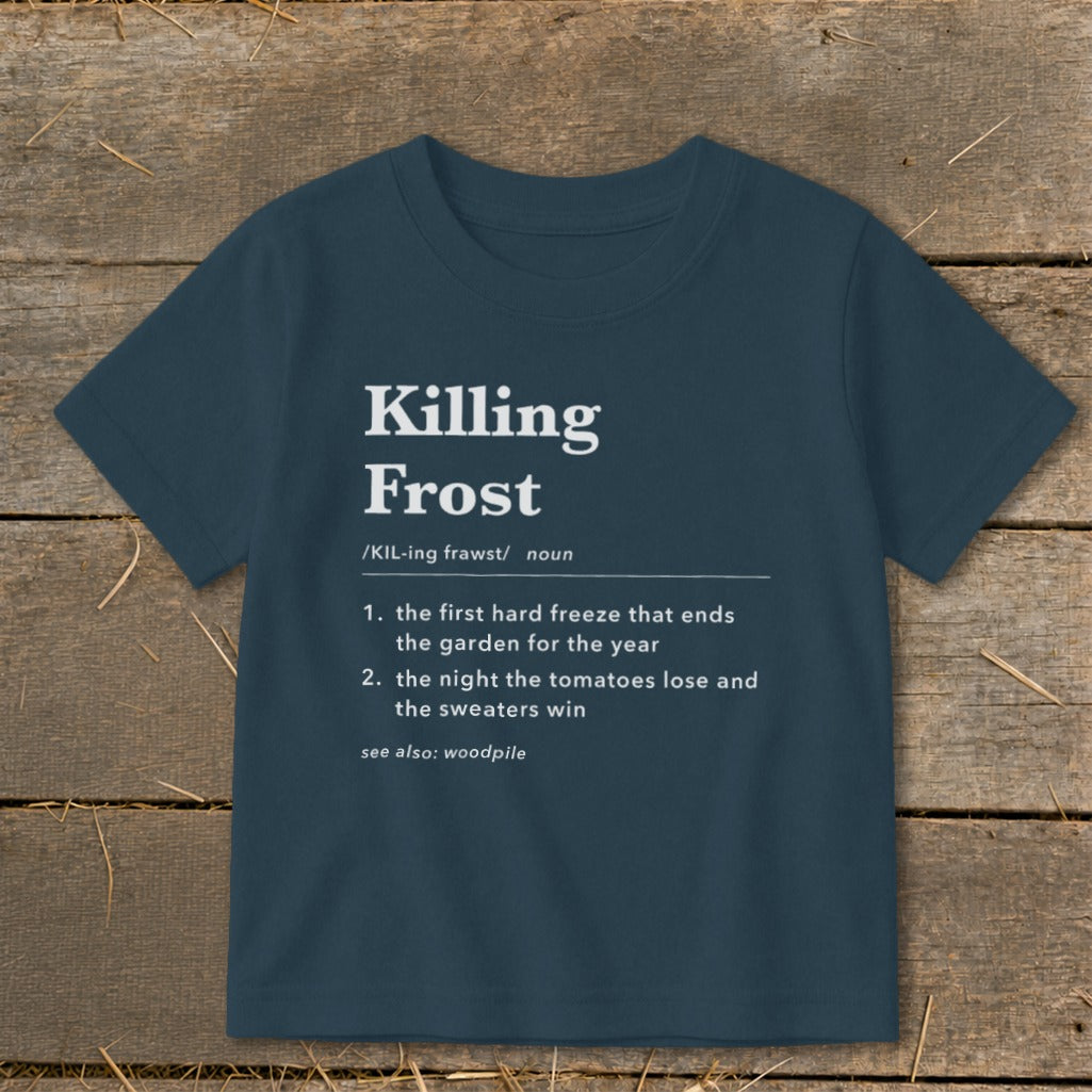 A dark blue short-sleeved t-shirt features white dictionary-style text defining killing frost laid flat on a rustic wooden background.