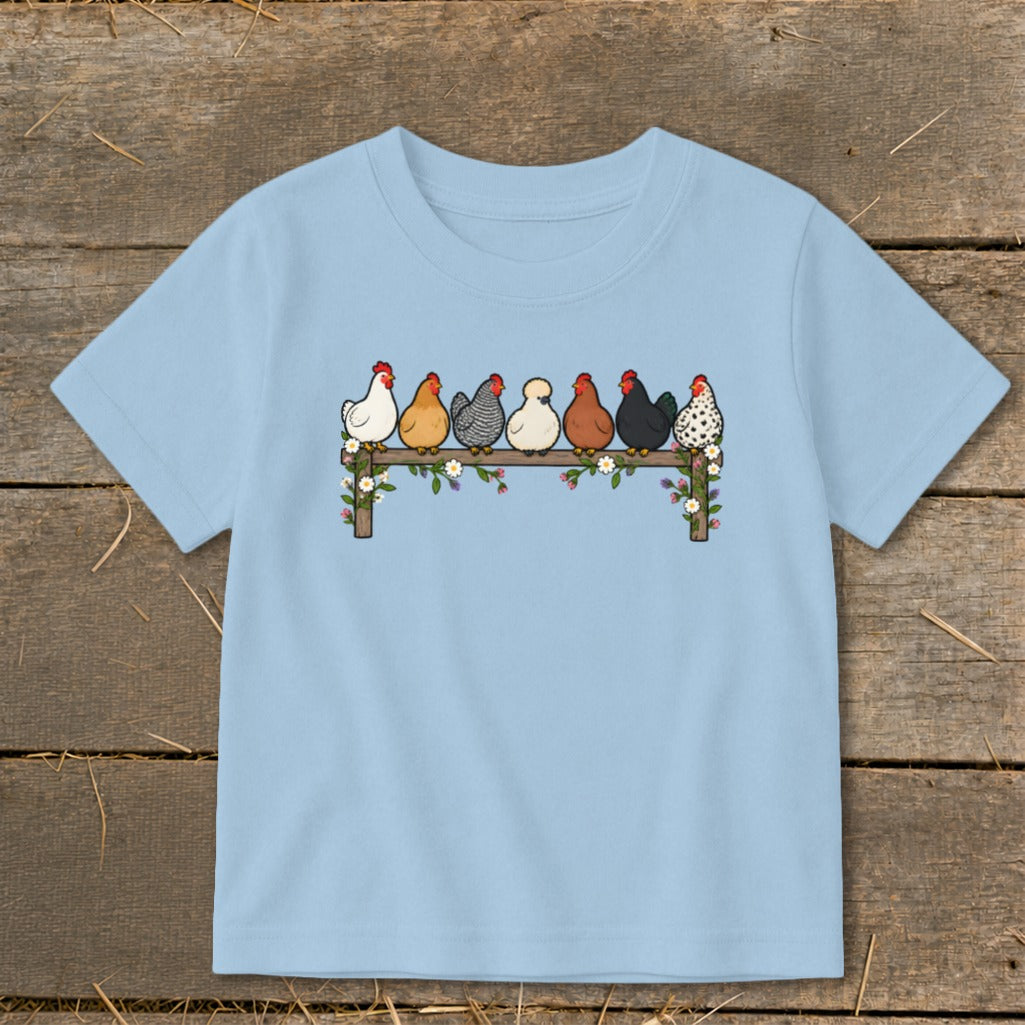 A light blue children's t-shirt features an illustration of seven colorful chickens perched on a flower-covered wooden fence.