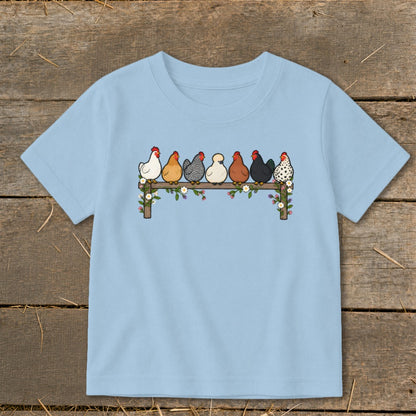 A light blue children's t-shirt features an illustration of seven colorful chickens perched on a flower-covered wooden fence.