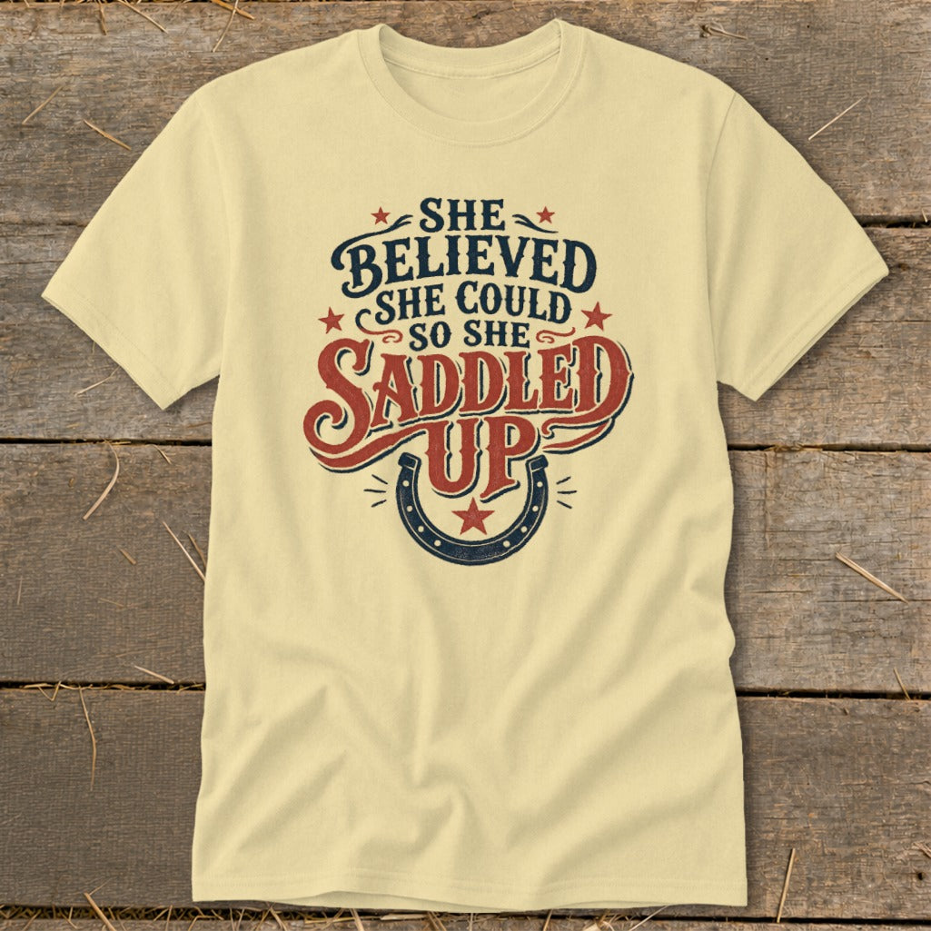 Pale yellow t-shirt with "She Believed She Could So She Saddled Up" text and horseshoe graphic.