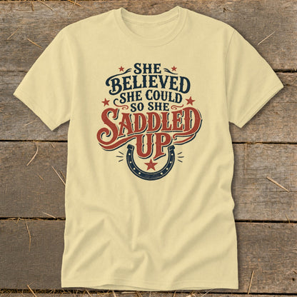 Pale yellow t-shirt with "She Believed She Could So She Saddled Up" text and horseshoe graphic.