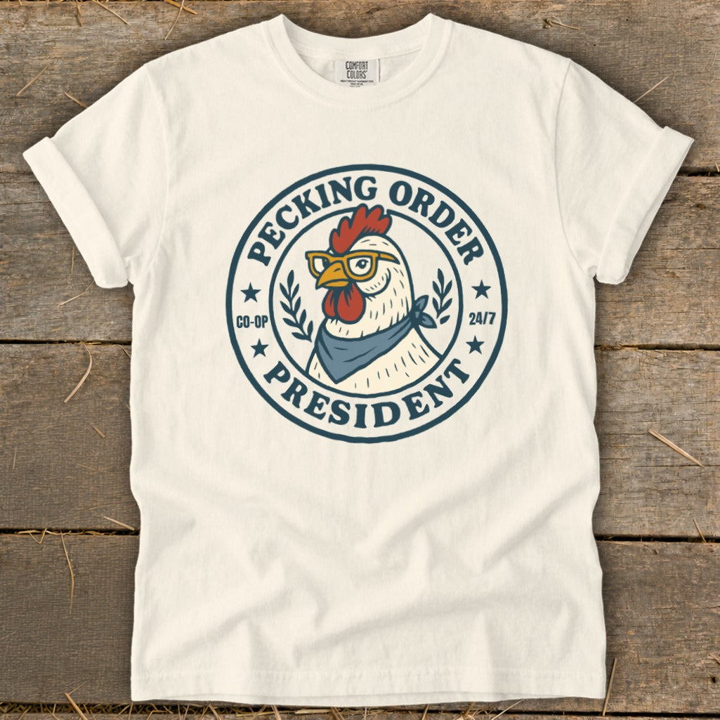 Cream t-shirt with a graphic of a chicken wearing glasses and a bandana, labeled "Pecking Order President Co-op 24/7".