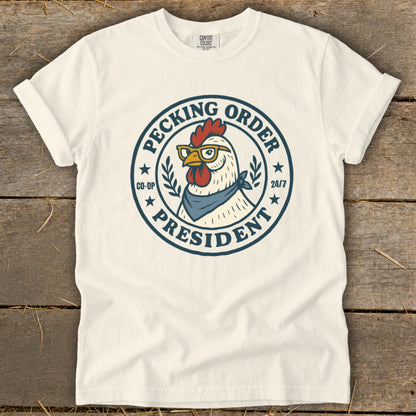 Cream t-shirt with a graphic of a chicken wearing glasses and a bandana, labeled "Pecking Order President Co-op 24/7".