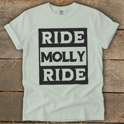 A light green t-shirt with rolled sleeves laid flat on a rustic wooden surface, featuring the black text "RIDE MOLLY RIDE" in a bold, blocky font on the chest.