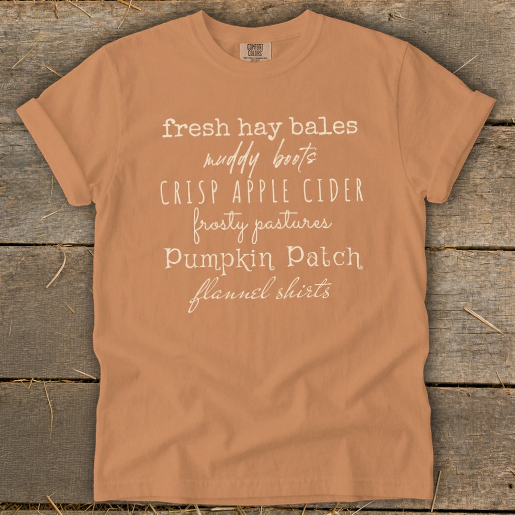 Orange t-shirt with white text listing fall phrases: fresh hay bales, muddy boots, crisp apple cider, frosty pastures, pumpkin patch, flannel shirts.