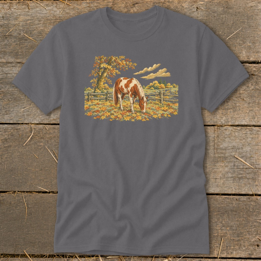 Brown and white horse grazing in a field of autumn leaves under a tree.