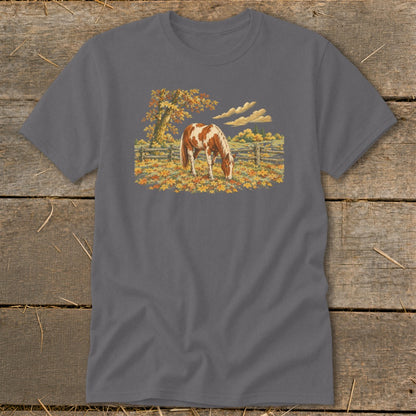 Brown and white horse grazing in a field of autumn leaves under a tree.