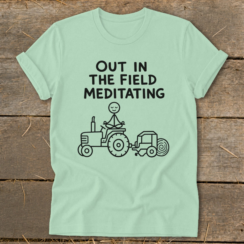 Light green t-shirt with text "OUT IN THE FIELD MEDITATING" and a drawing of a stick figure meditating on a tractor.