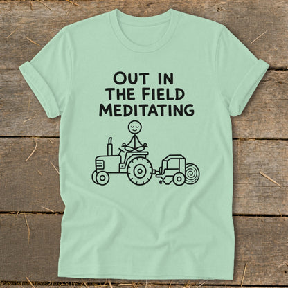 Light green t-shirt with text "OUT IN THE FIELD MEDITATING" and a drawing of a stick figure meditating on a tractor.