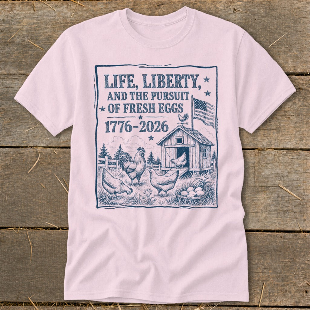 Pink t-shirt with a graphic of chickens, a coop, and the text "LIFE, LIBERTY, AND THE PURSUIT OF FRESH EGGS 1776-2026".