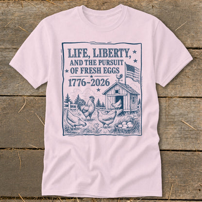 Pink t-shirt with a graphic of chickens, a coop, and the text "LIFE, LIBERTY, AND THE PURSUIT OF FRESH EGGS 1776-2026".