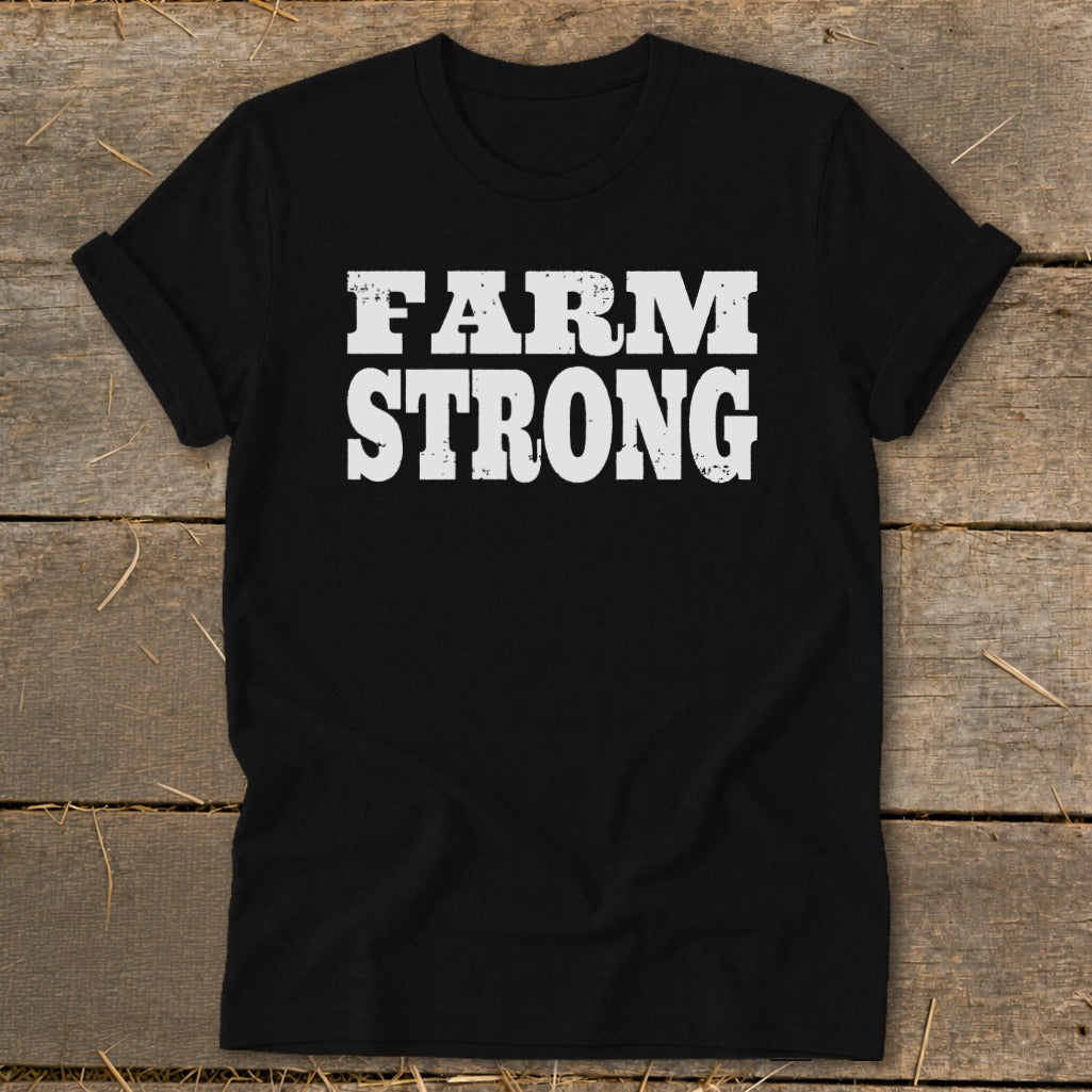 A black crewneck t-shirt with rolled sleeves displays the distressed white text FARM STRONG on a rustic wooden background.