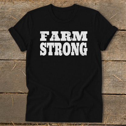 A black crewneck t-shirt with rolled sleeves displays the distressed white text FARM STRONG on a rustic wooden background.