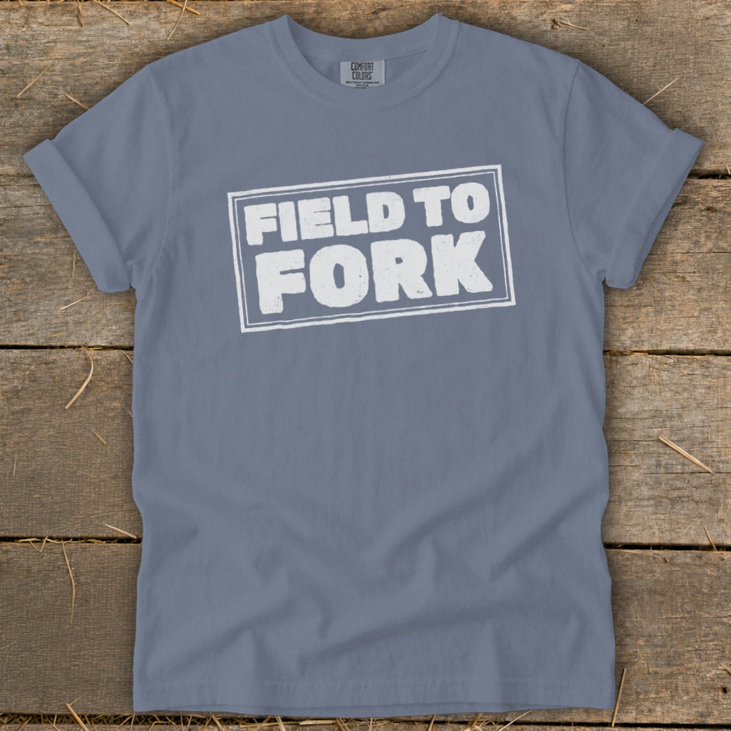 A blue-gray crewneck t-shirt with distressed white text reading FIELD TO FORK laid flat on a rustic wooden background.