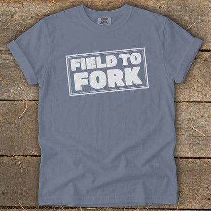 A blue-gray crewneck t-shirt with distressed white text reading FIELD TO FORK laid flat on a rustic wooden background.