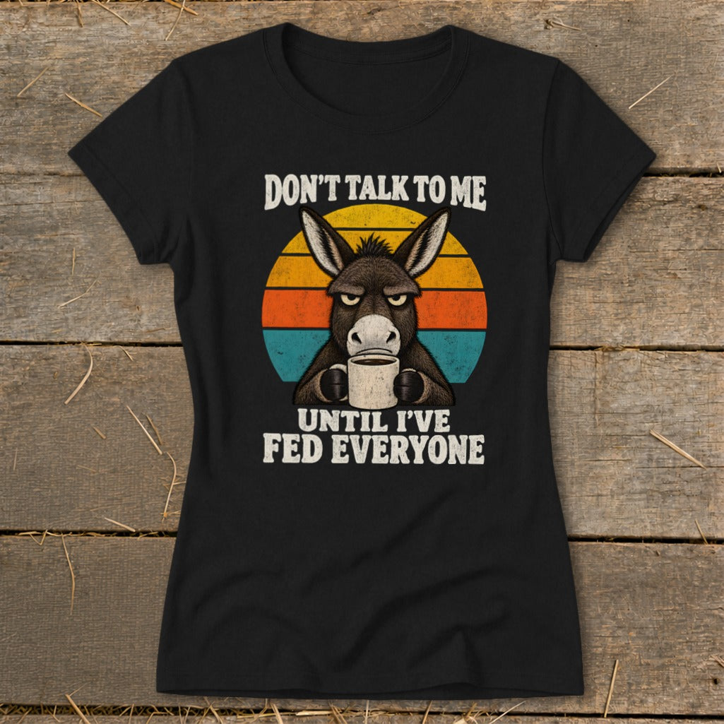 A black short-sleeved t-shirt featuring a graphic of an angry donkey holding a coffee mug against a retro sunset background, with the text DON'T TALK TO ME UNTIL I'VE FED EVERYONE.