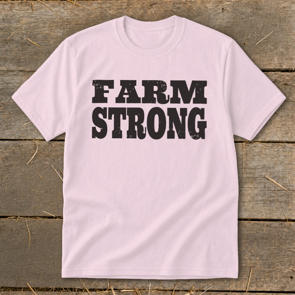 Light pink crew neck t-shirt with bold black distressed text that reads FARM STRONG, laid flat on a rustic wooden surface.