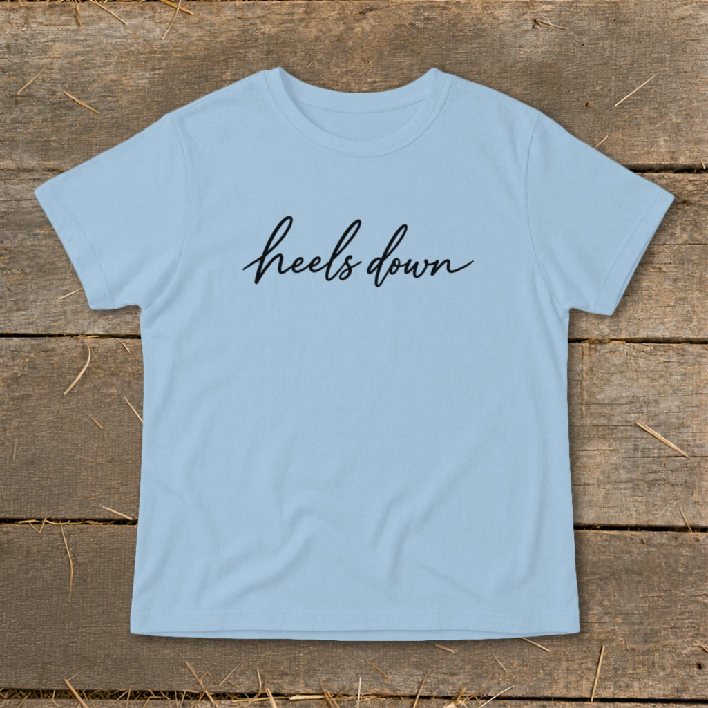 Light blue t-shirt with "heels down" text in black script on a wooden background.