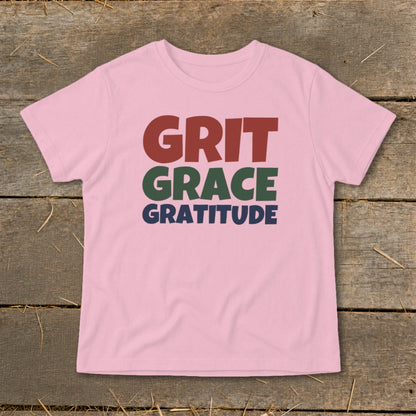 A pink t-shirt with the words "GRIT GRACE GRATITUDE" printed in red, green, and blue on a wooden background.
