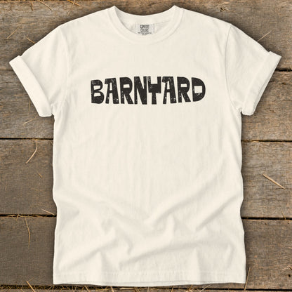 Off-white crewneck t-shirt featuring the black distressed text BARNYARD centered across the chest, laid on a rustic wooden surface.