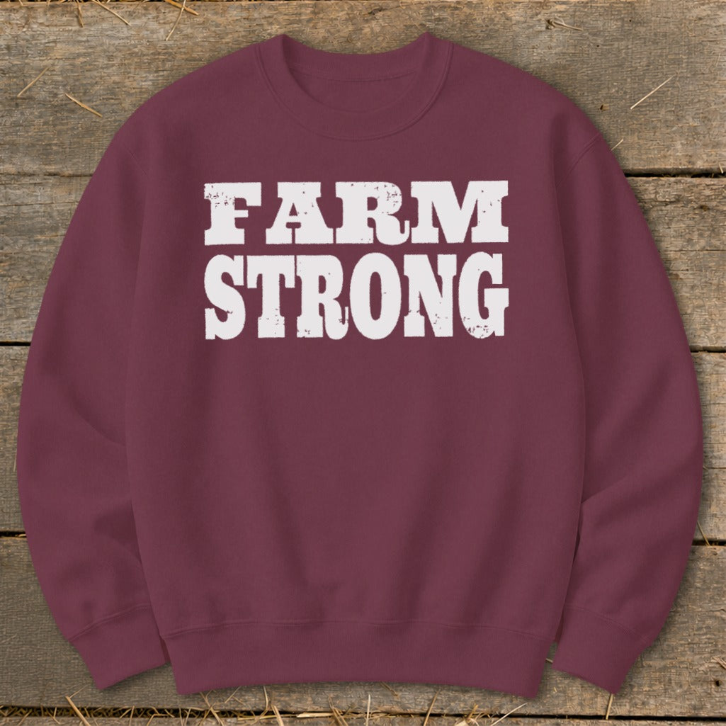 Maroon crewneck sweatshirt featuring the distressed white text FARM STRONG centered on the chest, laid flat on a rustic wooden background.