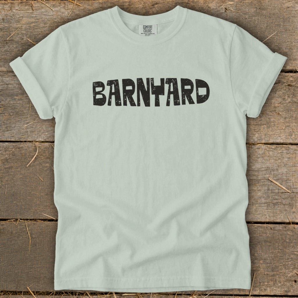A light green short-sleeve t-shirt with the word BARNYARD printed in distressed black font across the chest, laid flat on a rustic wooden background.