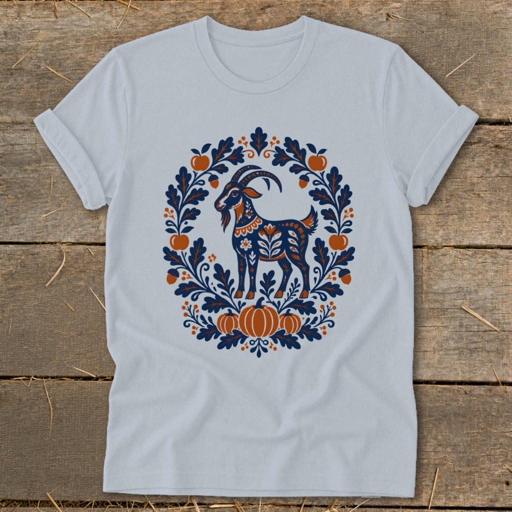 A light blue t-shirt displays an autumn-themed folk art graphic of a goat framed by leaves, acorns, and pumpkins.